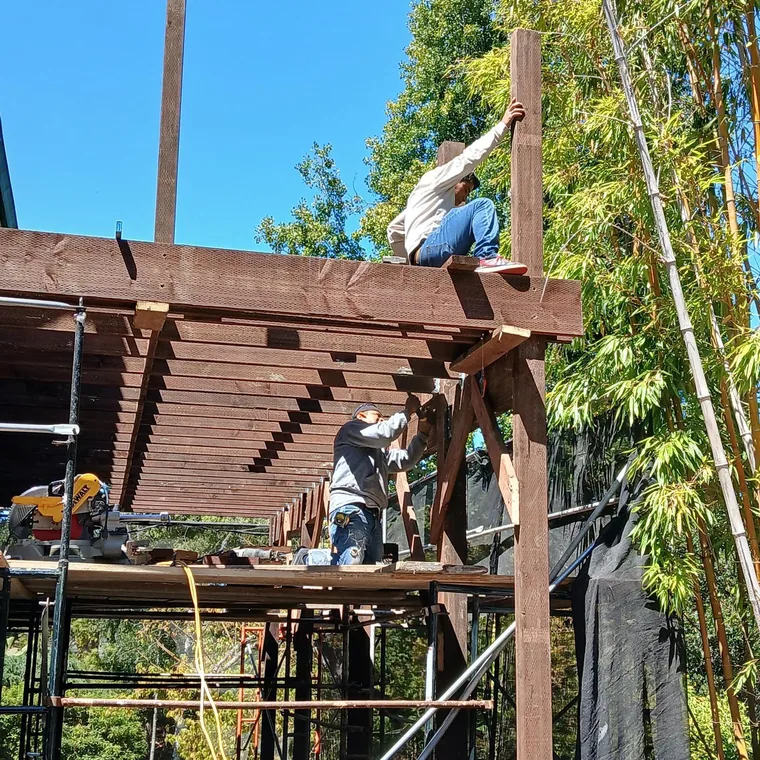 Two workers assembling elevated wood deck framing beside scaffolding