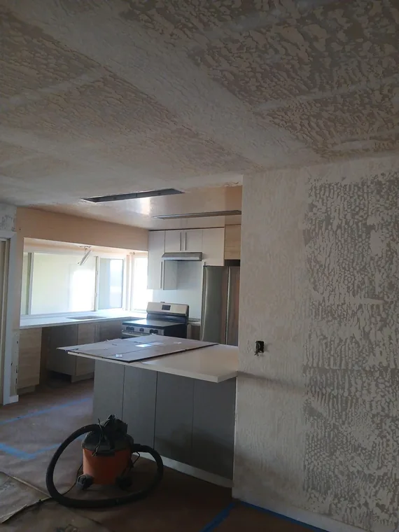 Wall and ceiling surfaces being prepared beside a kitchen, with a shop vacuum on the protected floor