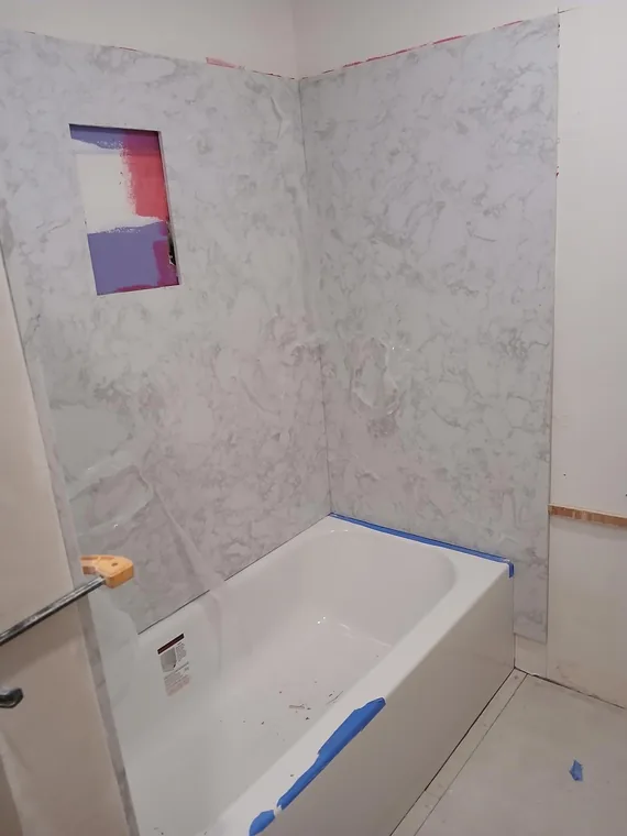White bathtub and marble-pattern wall panels with an unfinished niche and protective tape